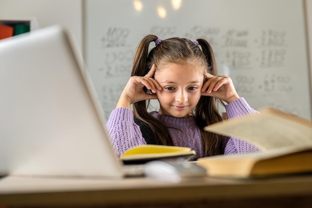 Cute little girl using laptop to study online at home pretty child sitting at the table looking at the computer screen and smiling enjoying distance learning close up