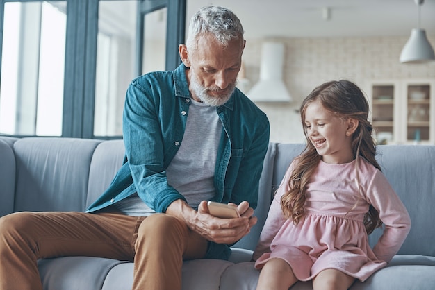 Cute little girl using smart phone while spending time with grandfather and at home