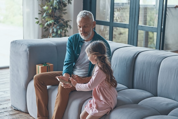 Cute little girl using smart phone while spending time with grandfather and at home