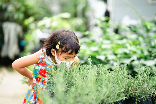 Cute little girl wearing protective face mask sniffing rosemary at garden