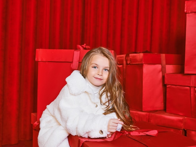 Cute little girl in white coat by the huge red christmas presents boxes on red background. Holidays