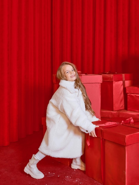 Cute little girl in white coat by the huge red christmas presents boxes on red background. Holidays