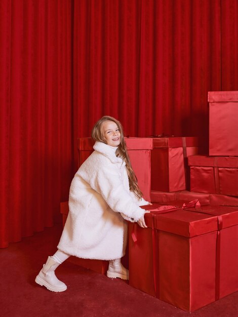 Cute little girl in white coat and had by the huge red christmas presents boxes on red background