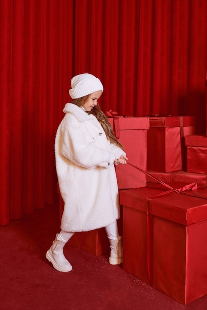 Cute little girl in white coat and had by the huge red christmas presents boxes on red background