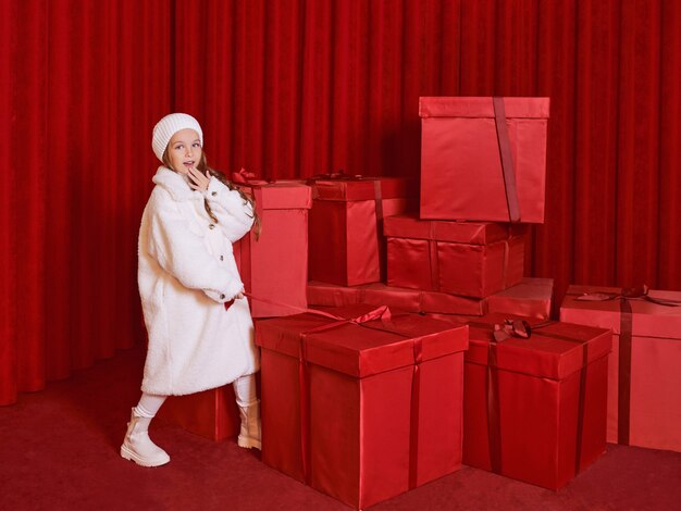 Cute little girl in white coat and had by the huge red christmas presents boxes on red background