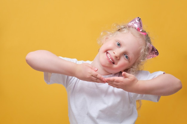 Cute little girl in a white tshirt having fun a child in a headband with cat ears on a yellow background