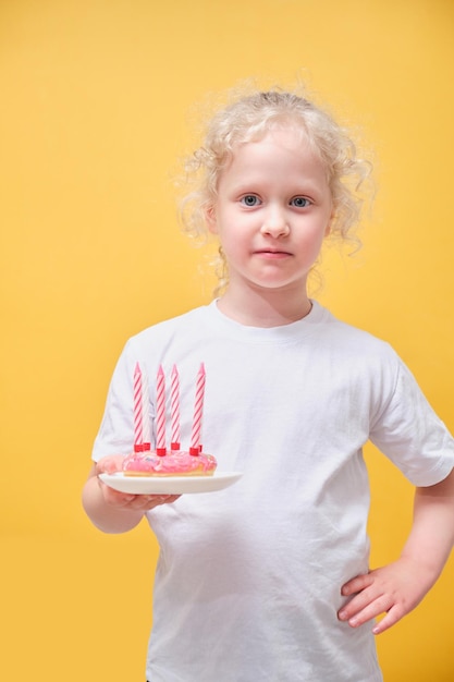 Cute little girl in a white tshirt holding a donut with candles having fun and making faces a child in a straw hat