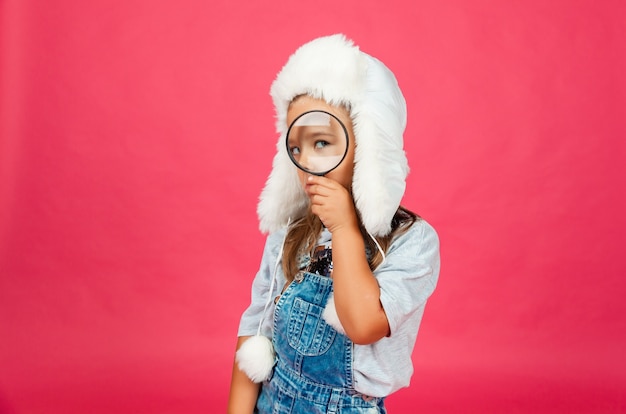 Cute little girl in a winter hat looking through a magnifying glass on a pink background.