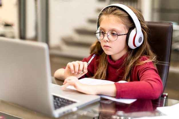 Cute little girl in wireless headphones seriously doing homework using laptop at home for online education and home studying. Kids distance learning. Social distance. Stay at home.