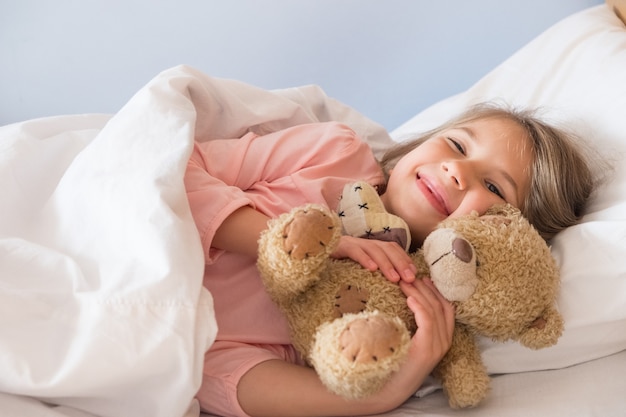 A cute little girl with blond hair in a pink tshirt lies in bed smiling and hugging a teddy bear