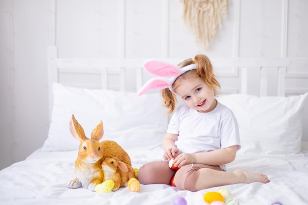 A cute little girl with bunny ears and colorful eggs on a white bed at home playing and smiling a blonde child celebrates happy Easter