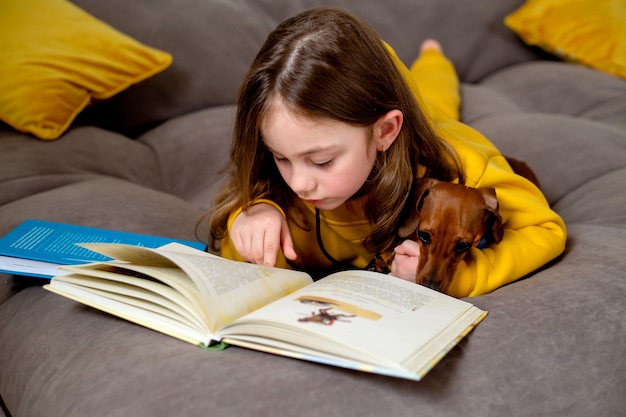 Cute little girl with a dog are lying on the bed and reading a book an open book is lying on bed