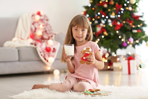 Cute little girl with glass of milk and cookie in room decorated for Christmas