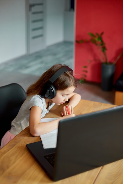 Cute little girl with headphones using laptop to study at home, writing, answer, online learning, education