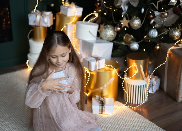 Cute little girl with long hair in dress sitting on the floor and opens a box with a present for background Christmas tree Xmas holiday concept