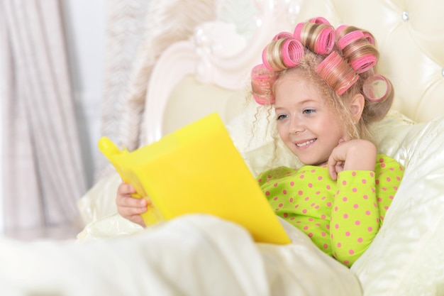 Cute little girl with pink hair curlers lying in bad and reading book