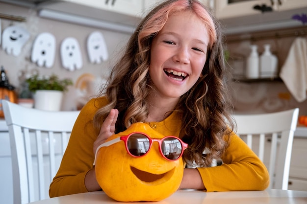 Cute little girl with pumpkin in sunglasses sitting in kitchen Halloween lifestyle kid portrait Halloween preparations