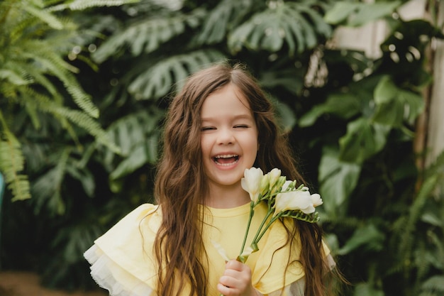 Cute little girl in a yellow dress holding spring flowers in her hands standing against the background of leaves