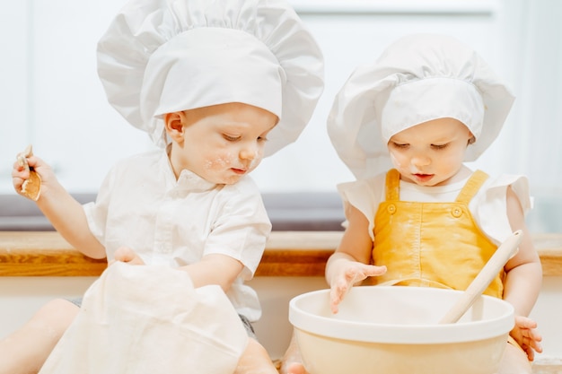 Cute little grimy caucasian children in hats are sitting on the table and stirring the dough for fritters in a bowl. The concept of busy skillful independent passionate children