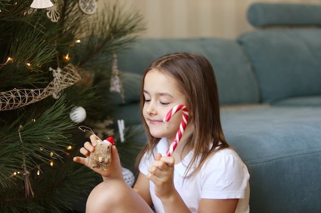 Cute little happy girl decorating Christmas tree holding striped candy cane and hedgehog t