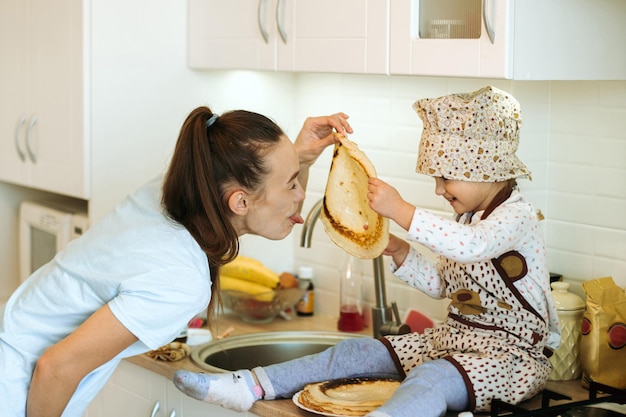 Cute little homecook girl with her beautiful mother makes pancakes in white kitchen