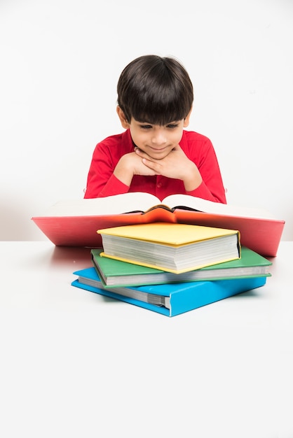 Cute little Indian or Asian boy holding or reading book over study table or over white floor, isolated over white background