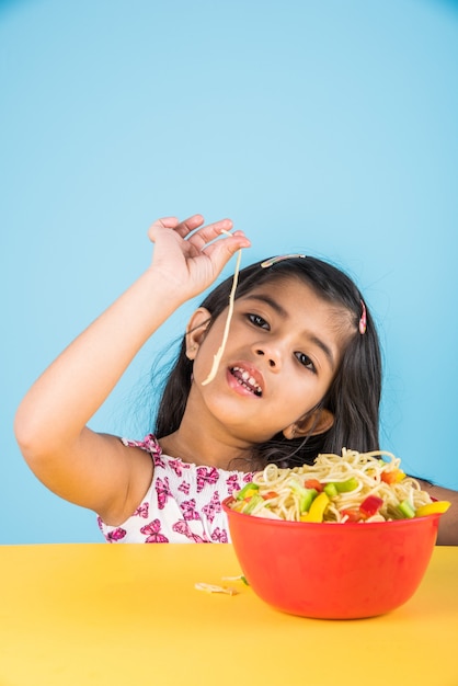 Cute little Indian or Asian girl child eating yummy Chinese Noodles with fork or chopsticks, isolated over colourful background