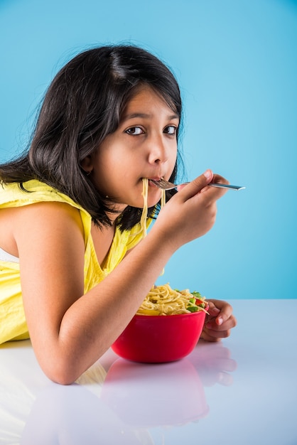 Cute little Indian or Asian girl child eating yummy Chinese Noodles with fork or chopsticks, isolated over colourful background