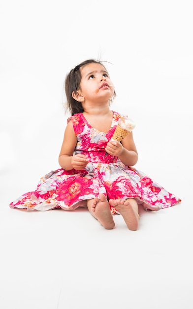 Cute little indian asian girl child licking or eating chocolate ice cream in cone, isolated over white background