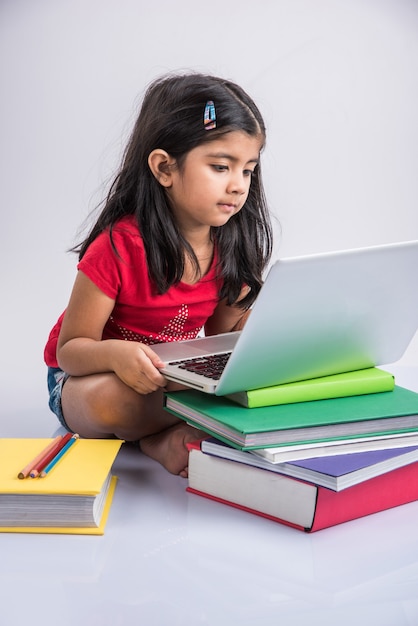 Cute little Indian or Asian girl child studying on laptop or working on school project while lying or sitting on the floor, isolated over white background