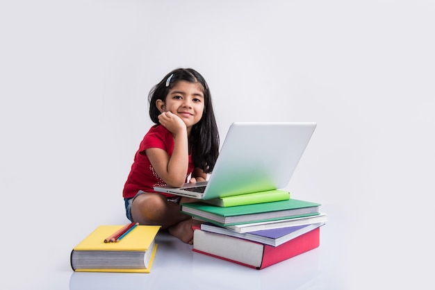 Cute little Indian or Asian girl child studying on laptop or working on school project while lying or sitting on the floor, isolated over white background