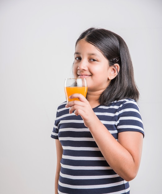 Cute little Indian or Asian playful girl drinking fresh mango or orange juice or cold drink or beverage in a glass, isolated over  white background