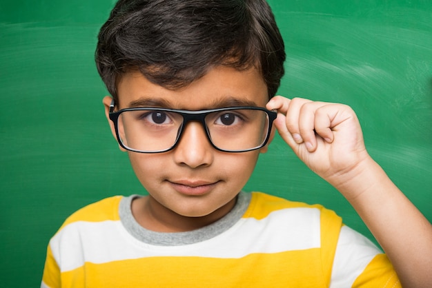 Cute little Indian school kid boy in hand stretched pose over green chalkboard or chalk board background holding books, victory cup etc, isolated