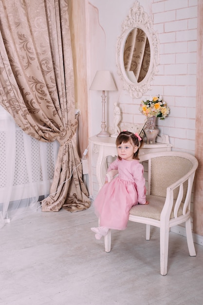 Cute little kid girl sitting on chair in pink dress at home