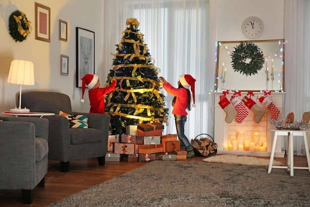 Cute little kids in Santa hats decorating Christmas tree at home