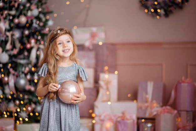 Cute little laughing blonde girl with long hair plays with a Christmas ball on a background