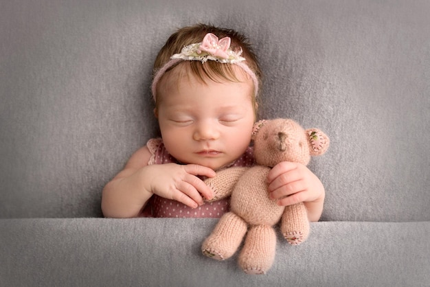 A cute little newborn baby in a pink suit and a pink headband with a butterfly on his head sleeps sweetly Knitted pear bear in hands Professional macro photo on a grey background