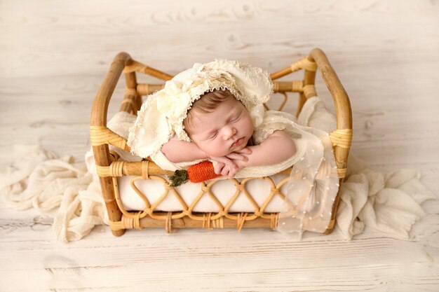 A cute little newborn baby in a white suit and a hat with bunny ears sleeps sweetly Wicker rattan bed with white linens In the hands of a red felt carrot Rabbit image light wood background