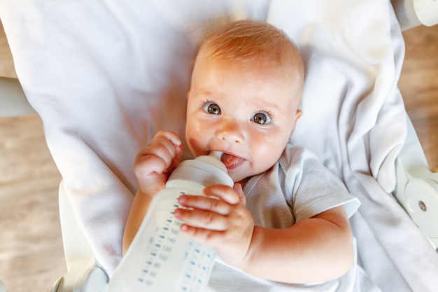 Cute little newborn girl drinking milk from bottle and looking at camera on white background. Infant baby sucking eating milk nutrition lying down on crib bed at home. Motherhood happy child concept.