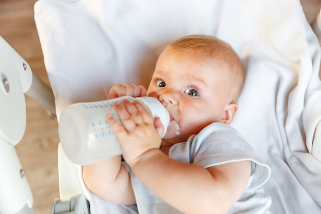 Cute little newborn girl drinking milk from bottle and looking at camera on white background. Infant baby sucking eating milk nutrition lying down on crib bed at home. Motherhood happy child concept.