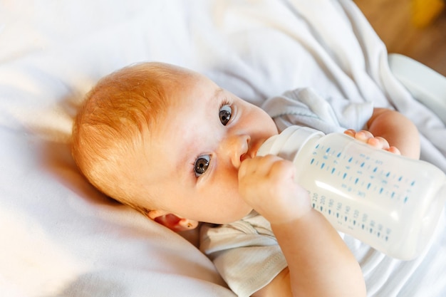 Cute little newborn girl drinking milk from bottle and looking at camera on white background Infant baby sucking eating milk nutrition lying down on crib bed at home Motherhood happy child concept