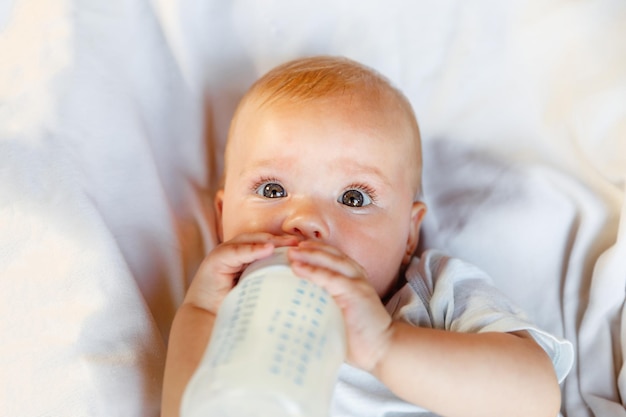 Cute little newborn girl drinking milk from bottle and looking at camera on white background Infant baby sucking eating milk nutrition lying down on crib bed at home Motherhood happy child concept