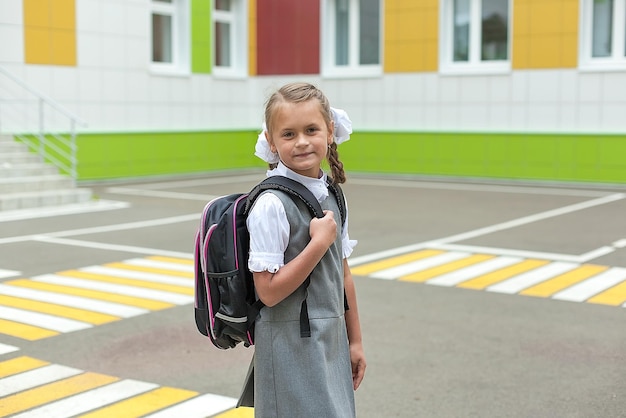 A cute little schoolgirl in a school uniform holds a backpack and smiles at the camera Return to school Knowledge Day