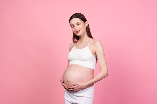 Cute lovely young caucasian woman touching her belly and looking tenderly straight, against a pink background. The concept of pregnancy, motherhood, preparation and expectation, firstborn. Copy space.