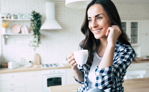 Cute lovely young woman in casual wear with coffee cup in hands is sitting on table in kitchen and relaxing