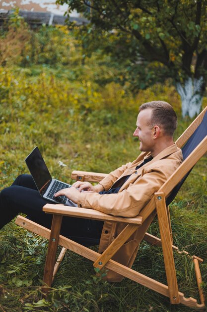 Cute man in a shirt sitting in a chair outdoors in the garden and working on a laptop remote work female freelancer