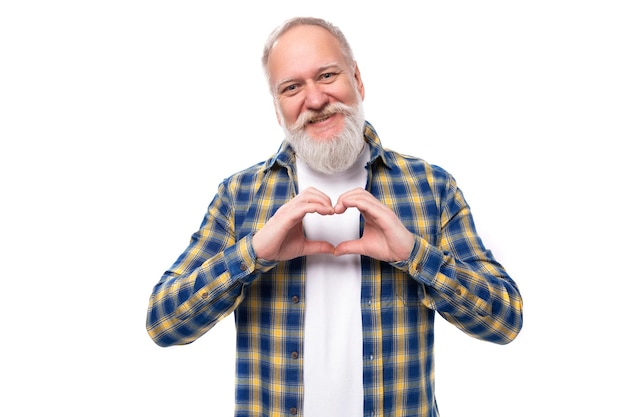 Cute mature grayhaired man with a beard in a shirt shows a heart with his hands on a white