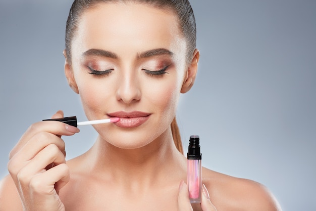 Cute model with brown hair fixed behind, clean fresh skin, big eyes and naked shoulders posing at gray studio background and looking at camera, close up, using lips gloss, closed eyes.