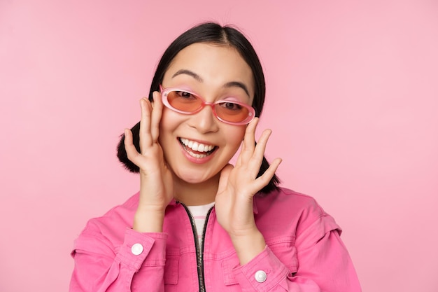 Cute modern japanese girl in sunglasses smiling and looking happy posing against pink background in stylish clothing