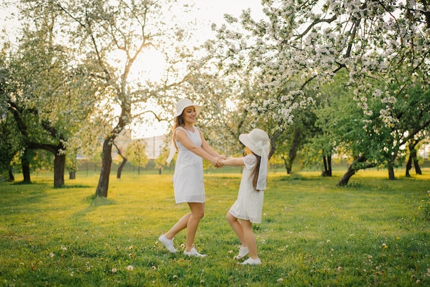 A cute mom in a hat and a white dress is walking through the spring garden with her cute daughter